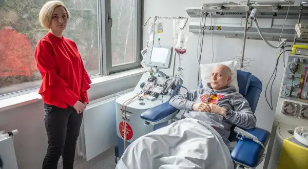 Kobieta w czerwonej bluzce stoi obok pacjenta podłączonego do aparatury medycznej. To ważny moment w procesie przeszczepu szpiku.