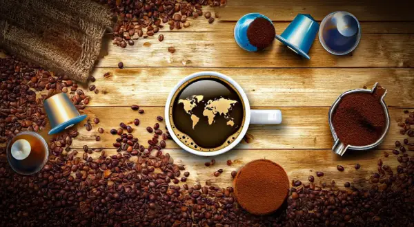 Tipi di cialde caffè e chicchi sparsi su un tavolo di legno. Una tazza di caffè con la mappa del mondo riflette il tema globale.