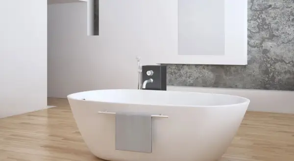 Eine freistehende **Badewanne mitten im Raum** auf hellem Holzboden, daneben ein moderner Wasserhahn und eine graue Handtuchhalterung.
