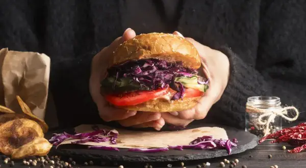 Ręce trzymają soczystego burgera z czerwoną kapustą, pomidorem i ogórkiem. Idealny przykład, jak składać burgera.