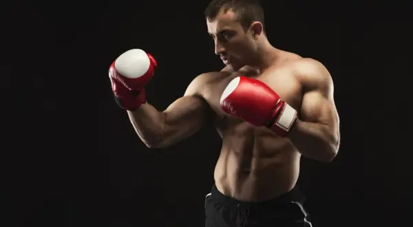 Muscular boxer in red gloves and black shorts in a defensive stance against a black background.