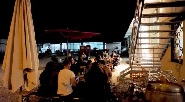 Serata al birrificio Mastro Matto: gente seduta ai tavoli sotto un grande ombrellone, con una scala metallica sullo sfondo.