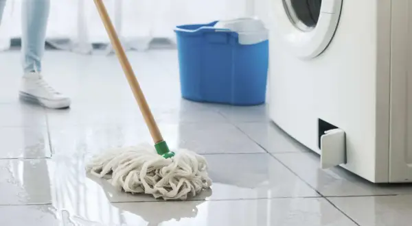 Eine Person wischt Wasser auf, das aus einer Waschmaschine austritt. Ein blauer Eimer steht bereit.