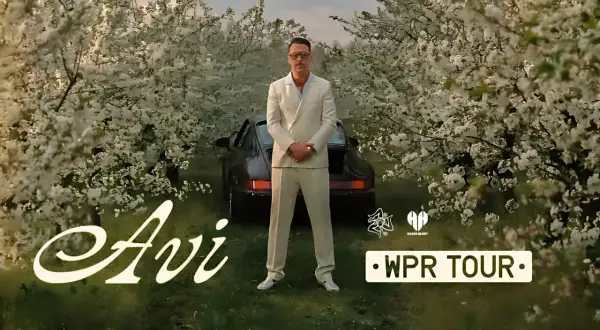 Mężczyzna w garniturze stoi przed czarnym samochodem wśród kwitnących drzew. Napis "Avi" i "WPR TOUR".
