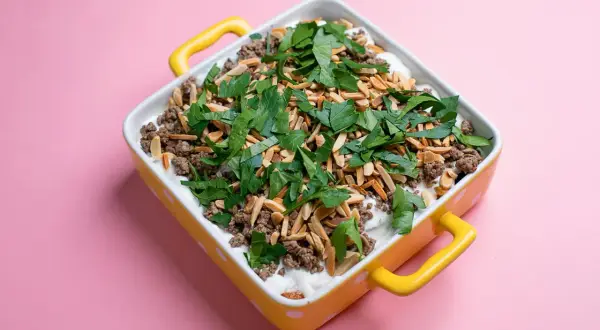 Un plat de fatteh aubergine garni de viande hachée, d'amandes effilées et de persil frais, dans un plat jaune à pois blancs.