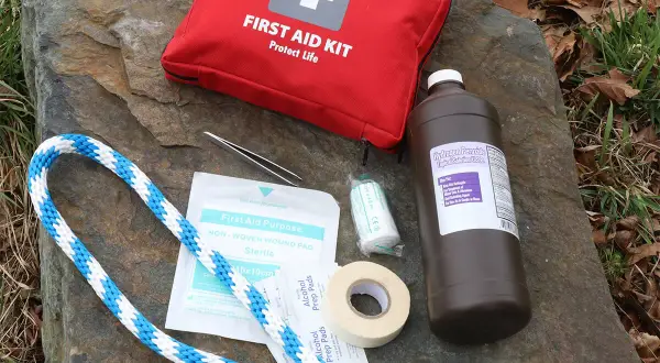 Contents of a DIY dog first aid kit laid out on a rock: red first aid bag, hydrogen peroxide, gauze, tape, alcohol pads, tweezers, and rope.