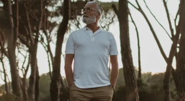 Älterer Mann mit Bart in weißem Polo und brauner Hose, steht im Wald. Die polo farben sind klassisch und zeitlos.