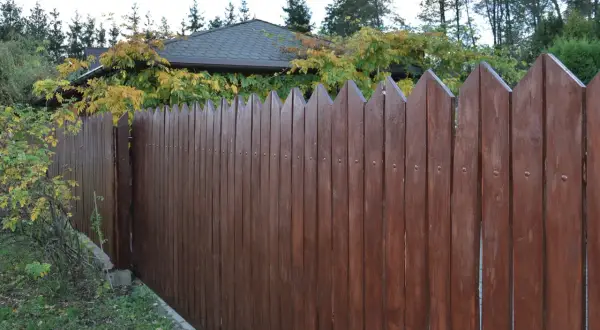Solidny płot z desek, pomalowany na ciemny brąz, chroni dom przed wzrokiem ciekawskich.