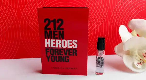 Czerwone opakowanie Carolina Herrera 212 Heroes Forever Young obok atomizera i białych kwiatów.