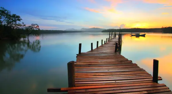 A wooden pier stretches into a calm lake at sunset, showcasing great depth of field photography. The sky transitions from blue to vibrant orange, reflecting on the water.