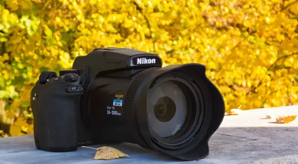 Czarny aparat Nikon P1000 z obiektywem na tle jesiennych, żółtych liści.
