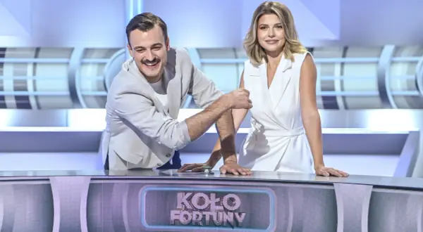 Uśmiechnięty mężczyzna i kobieta w białej sukience, którzy prowadzą Koło Fortuny, stoją przy pulpicie.