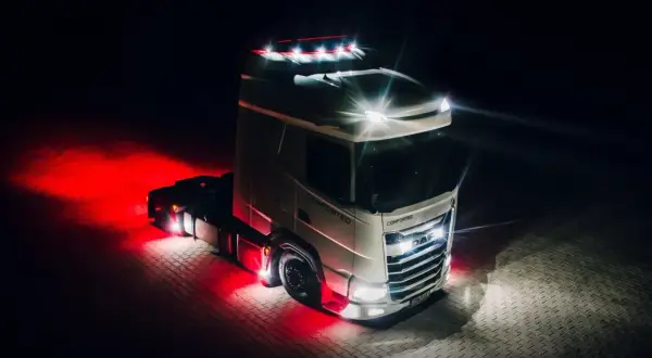 Camion DAF blanc illuminé par des feux de jour et des lumières rouges vives sur un sol pavé la nuit.