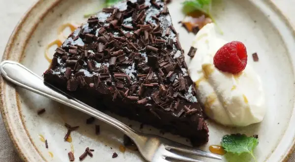 En bit glutenfri kladdkaka med chokladspån, vispad grädde, hallon och mynta på en tallrik.