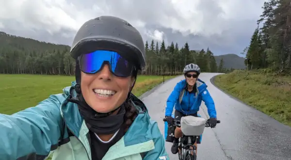 Dos ciclistas disfrutan de la ruta EuroVelo 6, con un paisaje verde y montañoso bajo un cielo nublado.