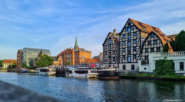Bydgoszcz atrakcje: łodzie cumujące przy Brdzie, nowoczesna architektura i zabytkowe kamienice z postacią na linie.