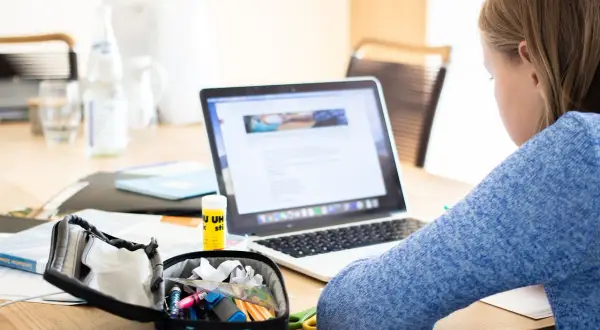 Dziewczynka uczy się przy laptopie, co pokazuje, czy nauczanie indywidualne może być prowadzone zdalnie. Na biurku przybory szkolne i butelka wody.