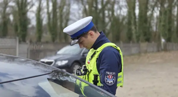 Policjant w kamizelce odblaskowej i czapce, pochylony nad maską samochodu, stosuje zasadę ograniczonego zaufania.