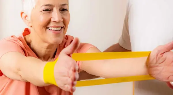Uśmiechnięta starsza kobieta ćwiczy z fizjoterapeutą, używając żółtej gumy rehabilitacyjnej.