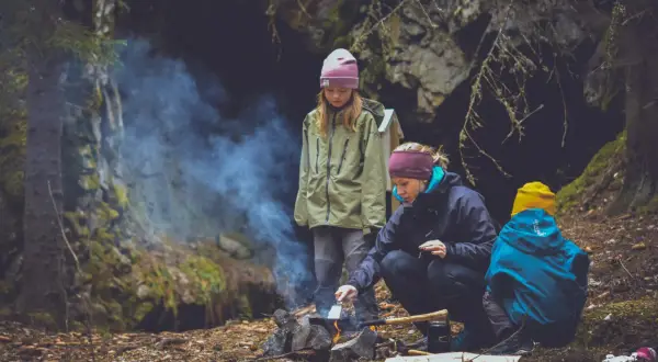 Familj lagar mat ute vid en lägereld i skogen. Rök stiger från elden där en kvinna steker något.