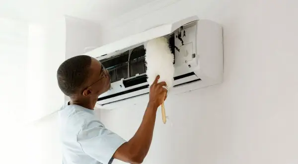 Un homme nettoie son climatiseur avec un plumeau. Un entretien régulier est essentiel pour savoir entretien climatisation tous les combien.
