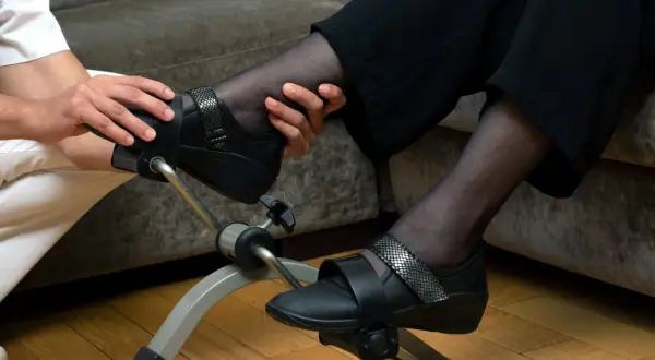 Osoba w białym stroju pomaga pacjentowi ćwiczyć na rowerku rehabilitacyjnym do wzmacniania mięśni rąk i nóg.