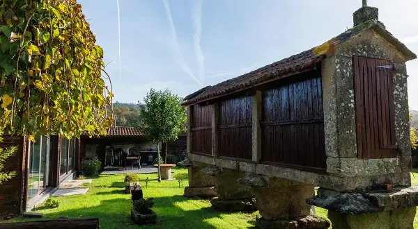 Un hórreo tradicional gallego, parte de la carballeira do Tambre, se alza sobre pilares de piedra. El sol ilumina el césped verde y un árbol frondoso.