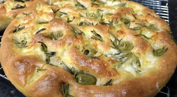 Une focaccia pizza dorée, garnie d'olives vertes et de romarin frais, sort du four.