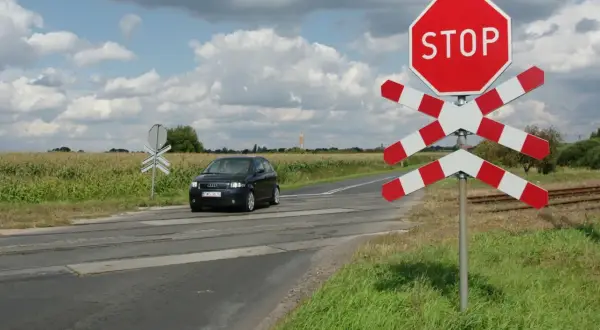 Przejazd kolejowy z zaporami i znakiem STOP. Samochód zbliża się do torów przez pole kukurydzy.
