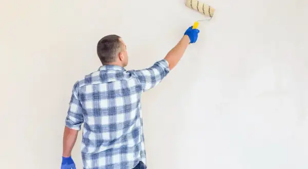 Mężczyzna w kratę nakłada wałkiem grunt pod tynk gipsowy na ścianę.