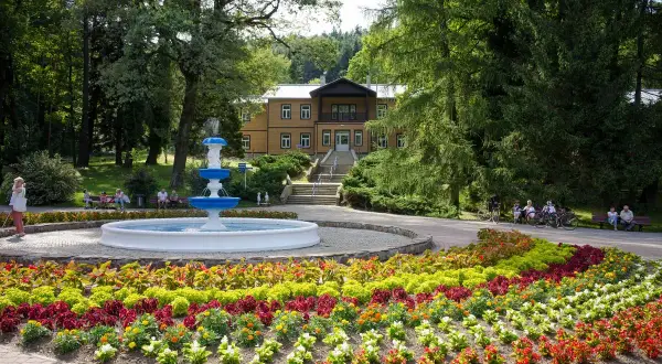 Uzdrowisko z fontanną i kolorowymi rabatami kwiatowymi przed budynkiem sanatorium.