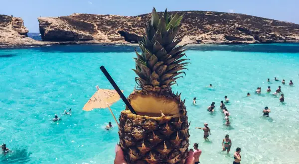 Koktajl z ananasa na plaży na Malcie. Woda jest turkusowa, a na horyzoncie widać skaliste klify. Ceny na Malcie są przystępne.