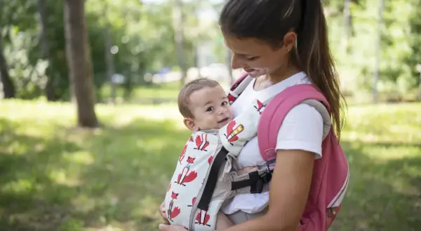 Uśmiechnięta mama z dzieckiem w nosidełku dla dziecka z lisami, na tle zielonego parku.