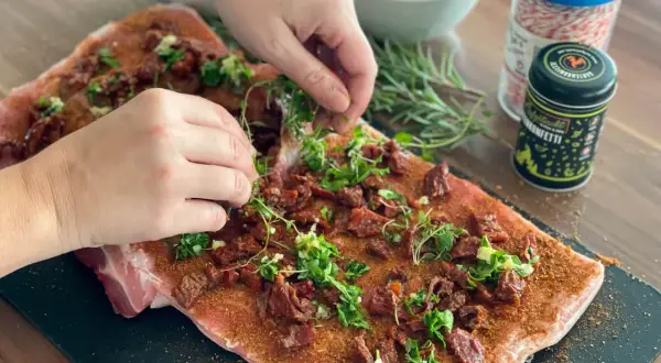 Hände belegen rohes Schweinefleisch mit Kräutern und getrockneten Tomaten für Porchetta.