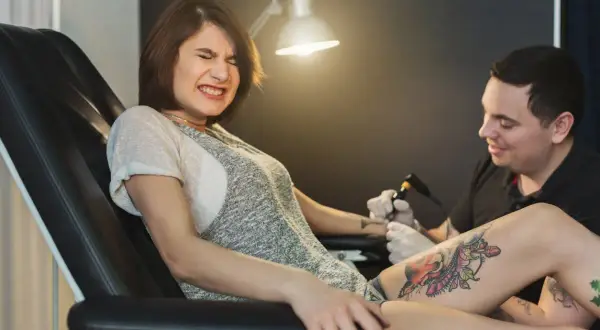 Mujer con expresión de dolor mientras le hacen un tatuaje en la pierna. El tatuador sonríe, ajeno al tattoo schmerzen.