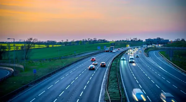 Autostrada z ruchem samochodowym o zachodzie słońca, z zielonymi polami w tle.