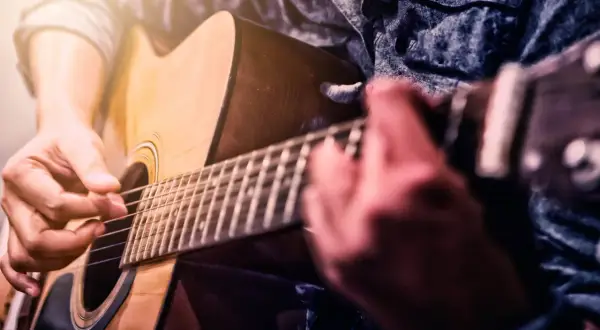 Ręce grające na gitarze akustycznej. Zbliżenie na palce naciskające struny i kostkę. Idealny moment, by zacząć naukę, jak nauczyć się grać na gitarze.