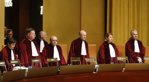 Siedmiu sędziów w togach, w tym jedna kobieta, stoi w sali sądowej. Wszyscy mają na sobie białe żaboty. W tle widać złote zasłony.