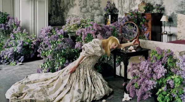 Una mujer rubia con un vestido estampado yace rodeada de lilas, una escena onírica capturada por Tim Walker.