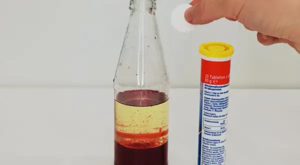 Eine Hand lässt eine Tablette in eine Flasche mit bunten Flüssigkeiten fallen, um eine Lavalampe selber zu machen.