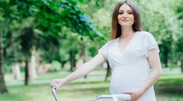 Uśmiechnięta ciężarna kobieta w białej sukience trzyma kierownicę roweru w parku. Czy w ciąży można jeździć na rowerze? Tak, jeśli czujesz się dobrze!