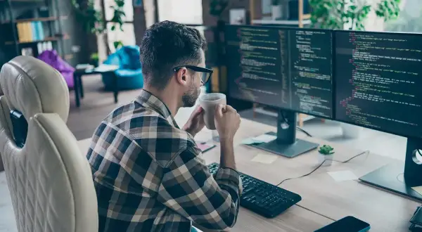 Programista w okularach pije kawę, pracując zdalnie przy dwóch monitorach z kodem.