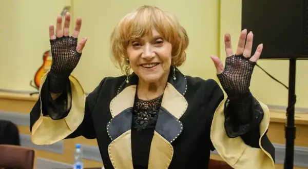 Uśmiechnięta starsza kobieta w czarnej marynarce z ozdobnym kołnierzem, w siatkowych rękawiczkach, unosi ręce.