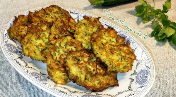 Frittelle di zucchine dorate e croccanti, servite su un piatto decorato. Accanto, una zucchina fresca e un rametto di erbe aromatiche.