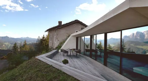 Modernes Haus in Hanglage mit Panoramafenstern und Terrasse, daneben ein altes Bauernhaus, umgeben von Berglandschaft.