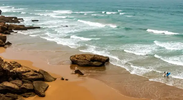 Skaliste marokańskie plaże z turkusową wodą i falami. Dwóch surferów wchodzi do oceanu z deskami.