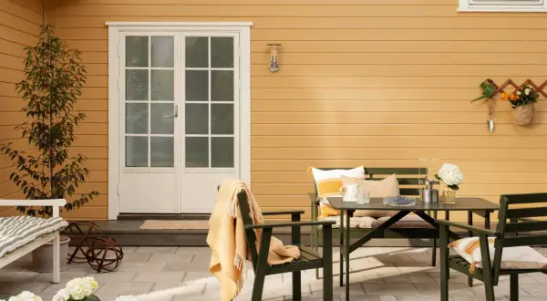 Ett gult hus med svart tak. En uteplats med gröna möbler, blommor och en orange pläd.