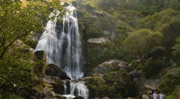 La majestuosa cascada de Belelle cae en cascada por rocas cubiertas de musgo, con gente disfrutando del paisaje y el agua.