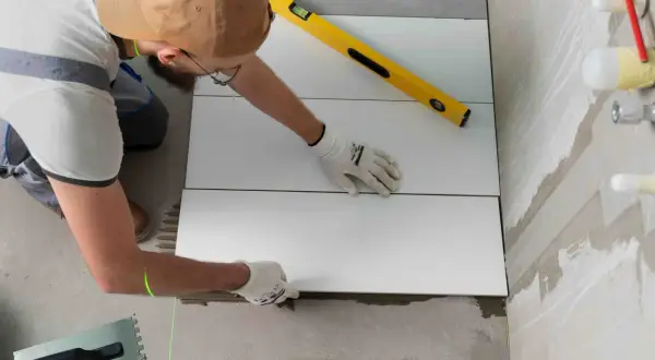 Un albañil coloca baldosas blancas sobre yeso, usando un nivel y un láser para asegurar una instalación perfecta.