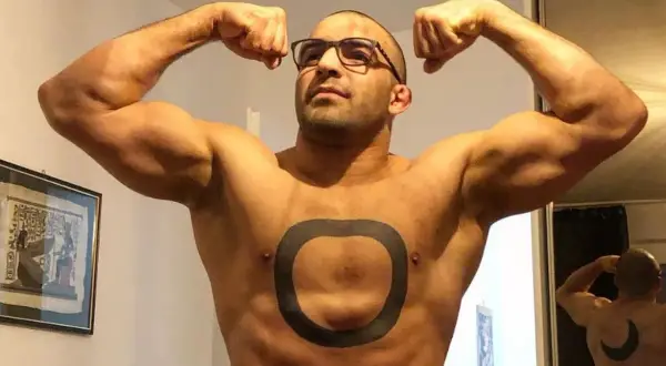 Muskularny mężczyzna o arabskich rysach, w okularach, prezentuje swoje bicepsy. Na klatce piersiowej ma tatuaż w kształcie pierścienia.
